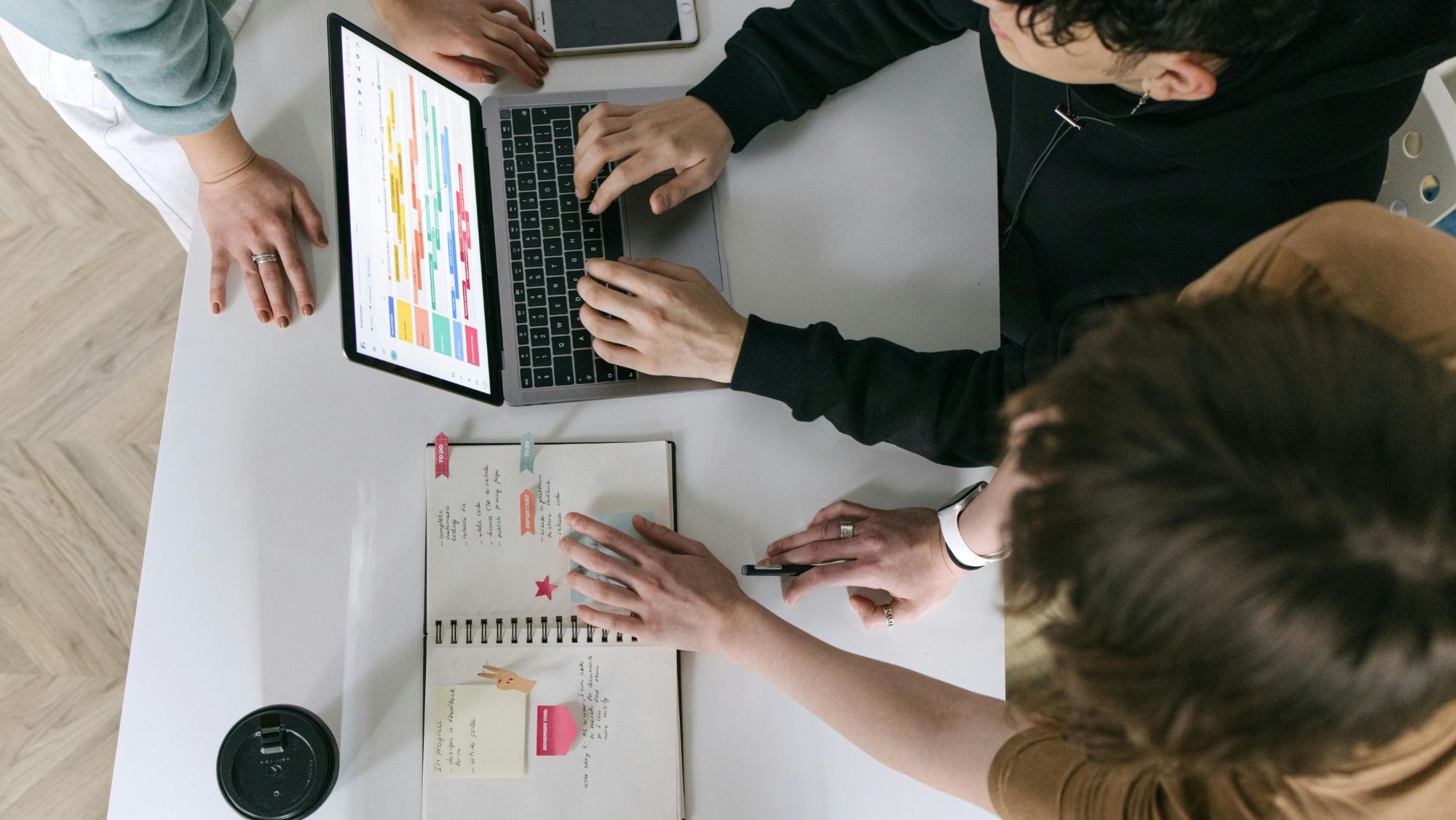 Två personer sitter vid ett bord och arbetar tillsammans. En öppen laptop med ett färgglatt schema syns, bredvid en anteckningsbok med handskrivna noteringar och post-it-lappar.